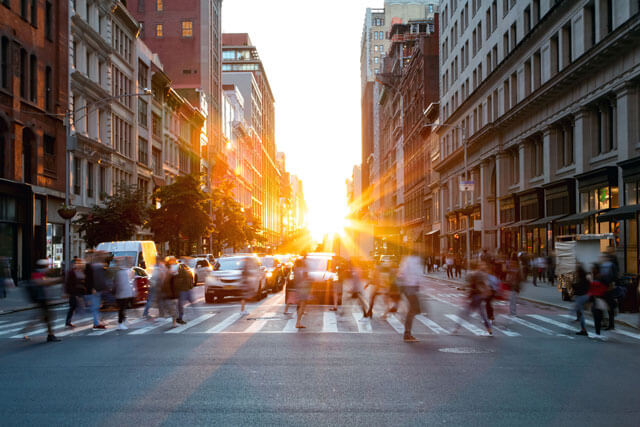 a street with sun setting down at background and people walking on a pedestrian crossing in the foreground